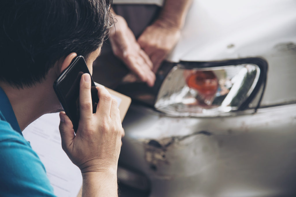 Foto de uma homem de camisa azul, segurando um celular preto enquanto olha para o farol esquerdo de um carro cinza danificado. Ao lado podemos perceber a mão de outra pessoa apontando para o dano. Imagem ilustrativa para texto proteção veicular. 