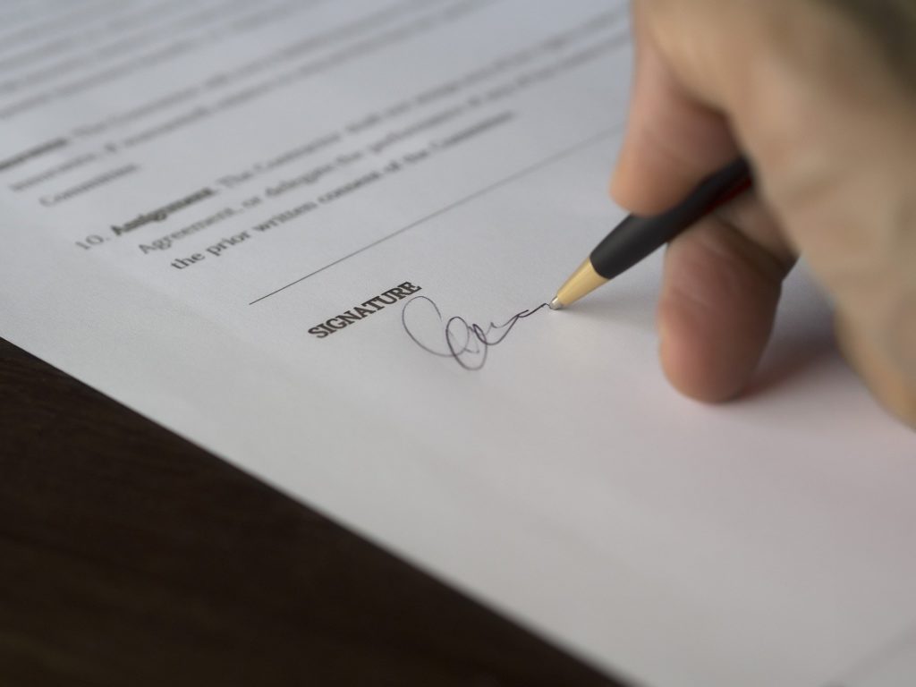 Foto de uma mão assinando um contrato com caneta preta. Vemos uma mesa de madeira. 