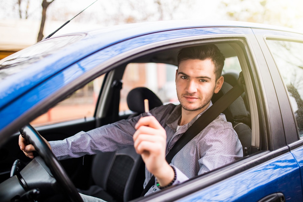 Homem de blusa cinza segurando a chave de um carro, enquanto está no banco do motorista. Vemos partes de um carro azul e uma paisagem desfocada ao fundo. 