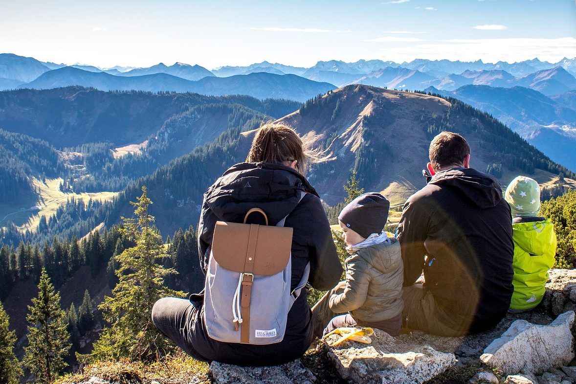 Foto de uma casal de roupa preta, a mulher usa mochila cinza, com duas crianças, uma de roupa cinza e outra de roupa verde. Todos estão sentados no topo de uma montanha, ao longe vemos uma paisagem. 