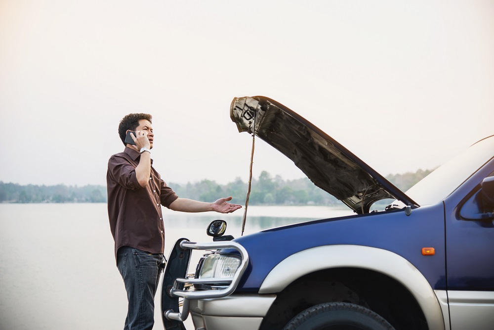 Homem de blusa marrom e calça jeans, fazendo uma ligação em frente a um carro azul estragado com o capô levantado. Ao fundo temos um lago e árvores.