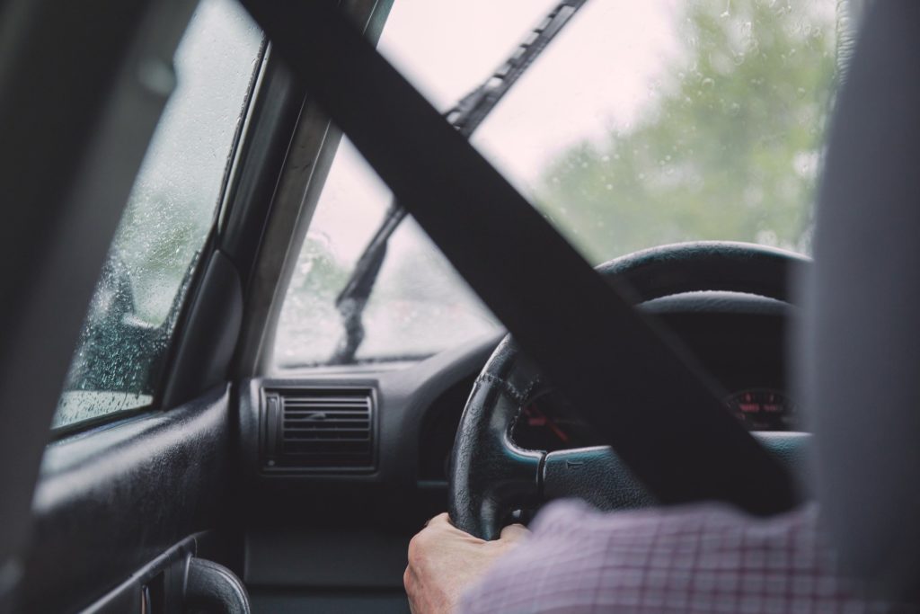 Foto de uma pessoa de roupa xadrez dirigindo um carro com o interior todo preto. Na janela vemos a chuva e uma paisagem de plantas. Imagem ilustrativa para texto seguros obrigatórios.